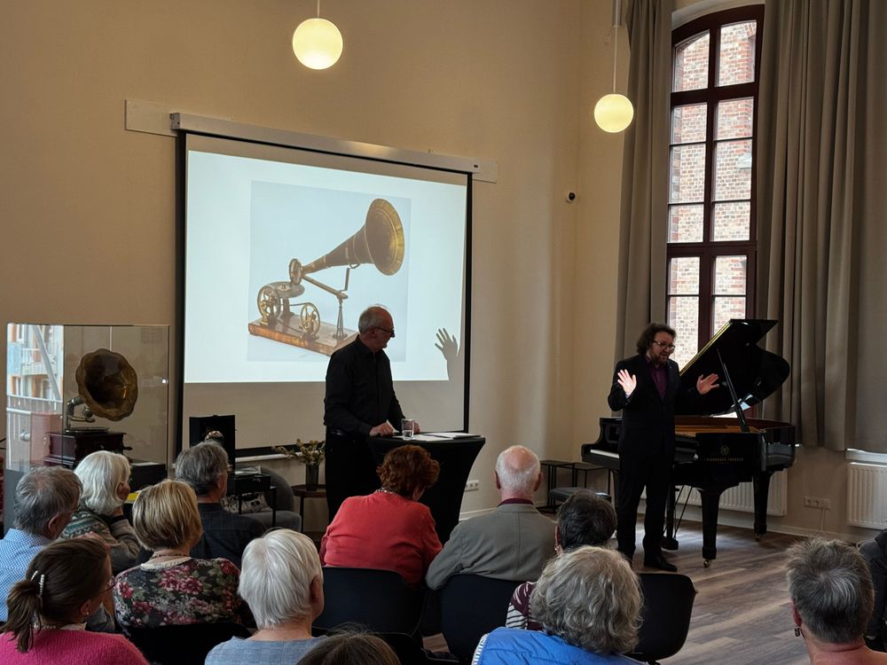 Der litauische Pianist Ginteras Janusevicius (rechts) und Referent Uwe Helms (links) gestalteten am 20. Mai 2026 im Emil Berliner Saal der ehemaligen Samsonschule in Wolfenbüttel einen Abend zum 175. Geburtstag des Erfinders und ehemaligen Samsonschülers Emil Berliner. Während Helms die Geschichte von Grammophon und Schallplatte erläuterte, begleitete Janusevicius den Vortrag mit virtuosen Klavierstücken. Im Hintergrund ist auf der Leinwand ein historisches Grammophon aus der Präsentation zu sehen, im Vordergrund verfolgt das Publikum aufmerksam die Veranstaltung