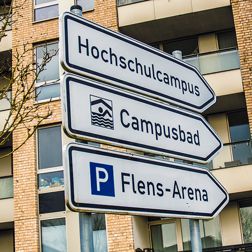 Ein Straßenschild weist zum Hochschulcampus, zum Campusbad und zur Flens-Arena in Flesnburg. Im Hintergrund ist ein mehrgeschossiges Neubauwohnhaus zu sehen. 